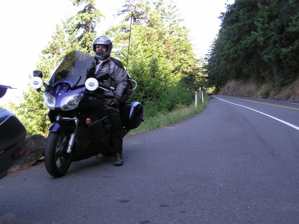 A rider on the blue 2005 FJR1300 with Hella driving lights, stopped on a two-lane road through the trees.